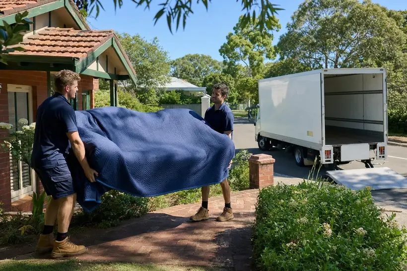 Two Perth removalists carrying a wrapped lounge from an established brick-and-tile family home on a leafy Cannington street with the plain removals truck at the kerb