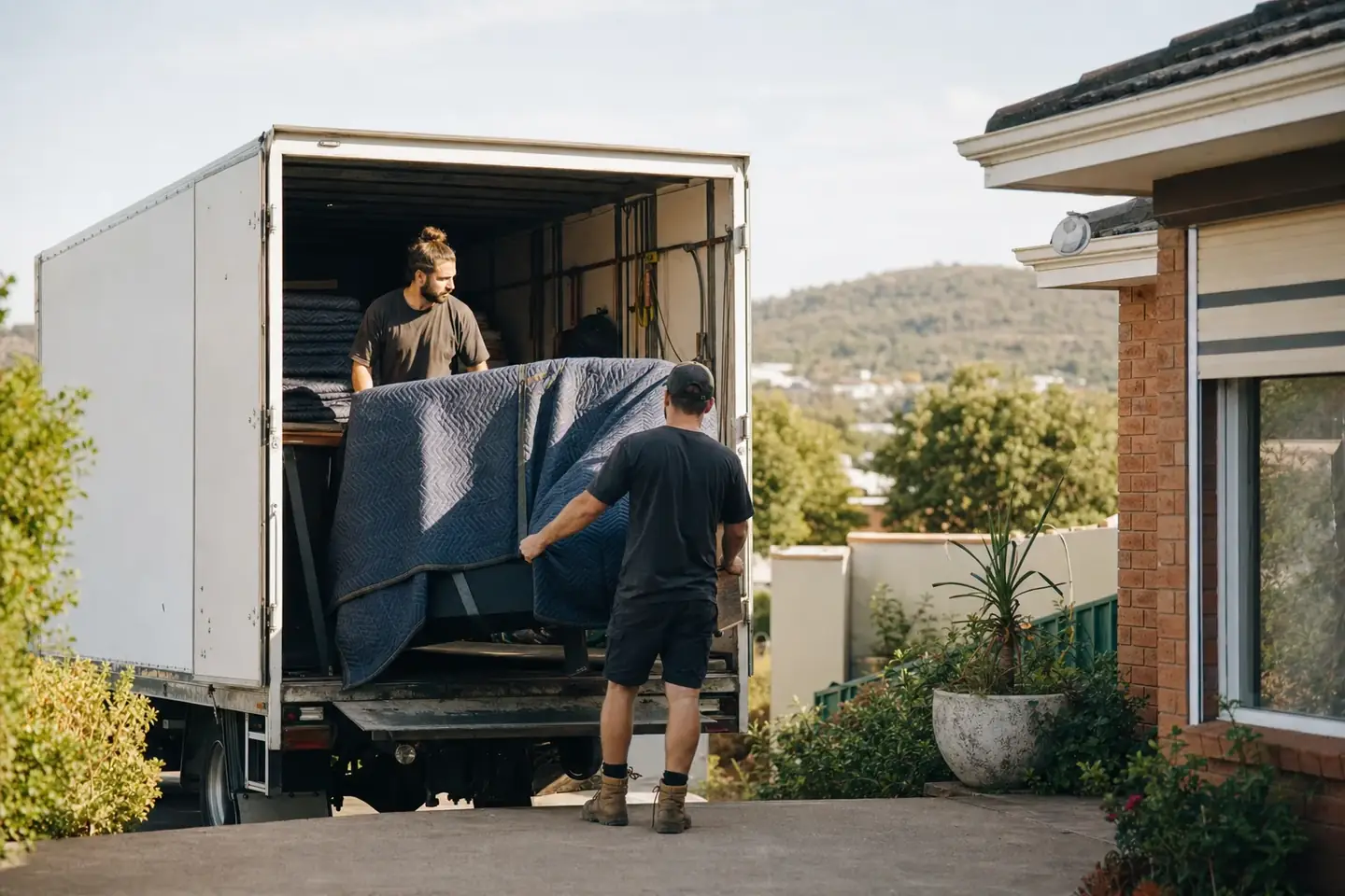 Two Perth removalists loading wrapped furniture into a truck outside a brick home in Midland with the green hills in the background