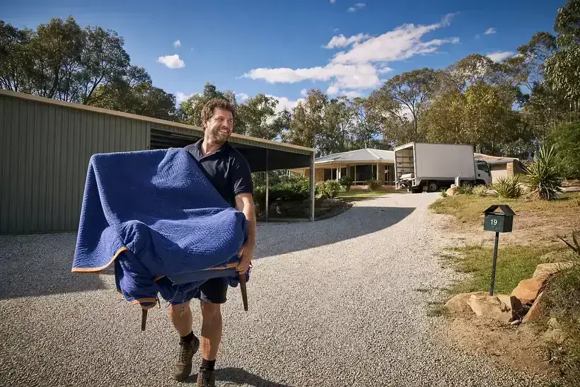 Perth removalist carrying a wrapped armchair across a large semi-rural Wanneroo property with a big shed and the removals truck near the house