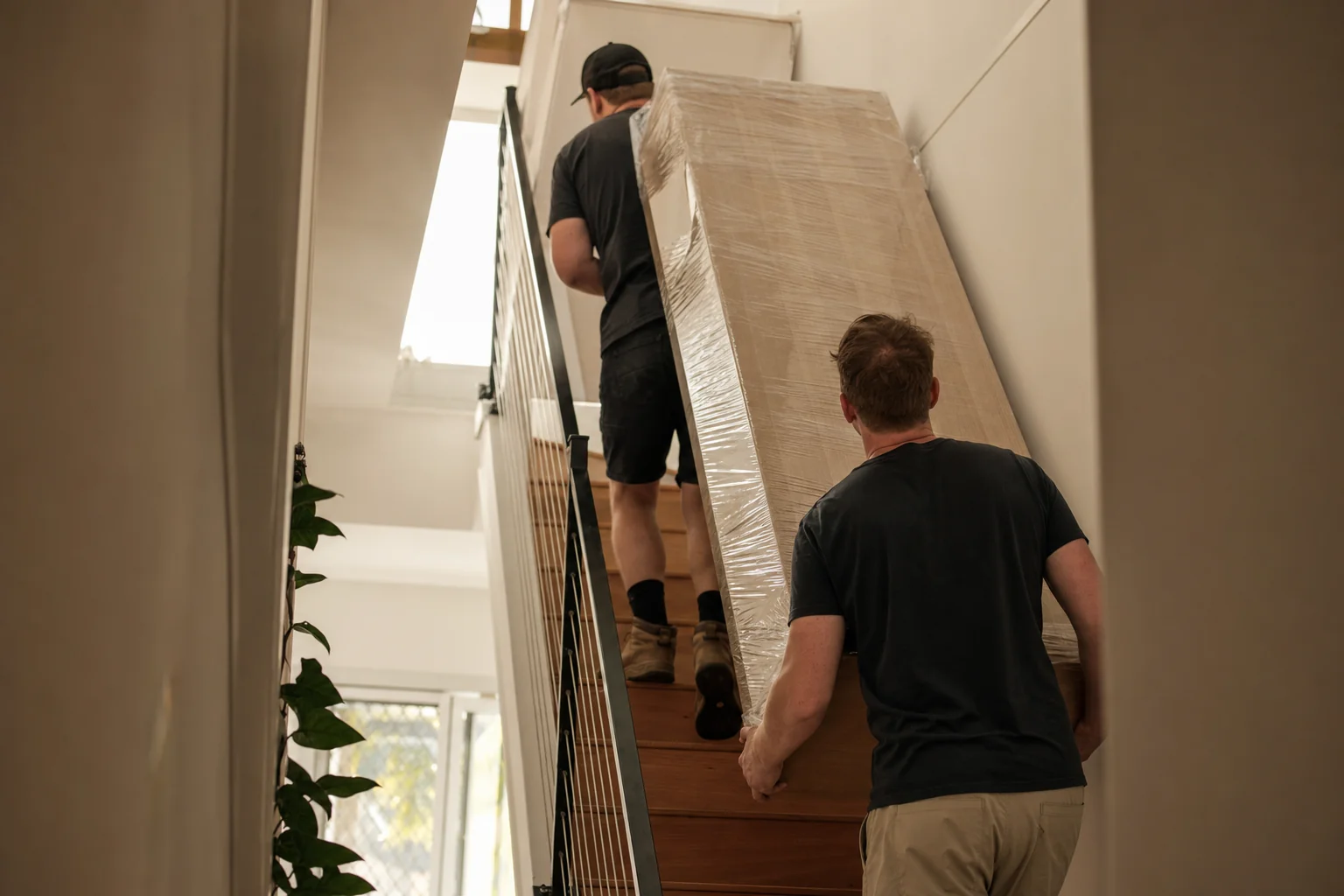 Two Perth removalists carrying a wrapped flat-pack wardrobe up the internal staircase of a modern double-storey Wellard home