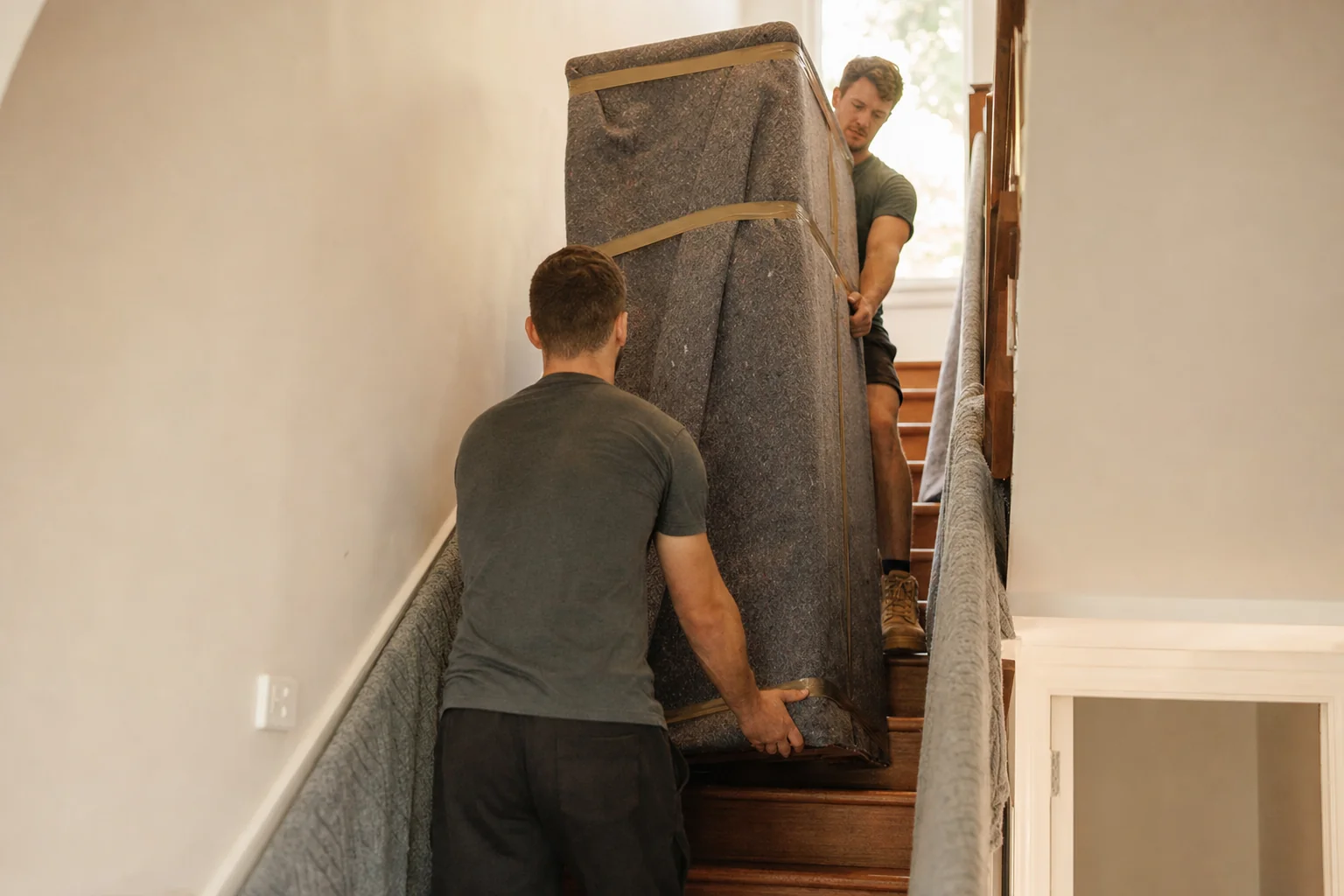 Two Perth removalists carrying a wrapped wardrobe up a narrow staircase with a spotter