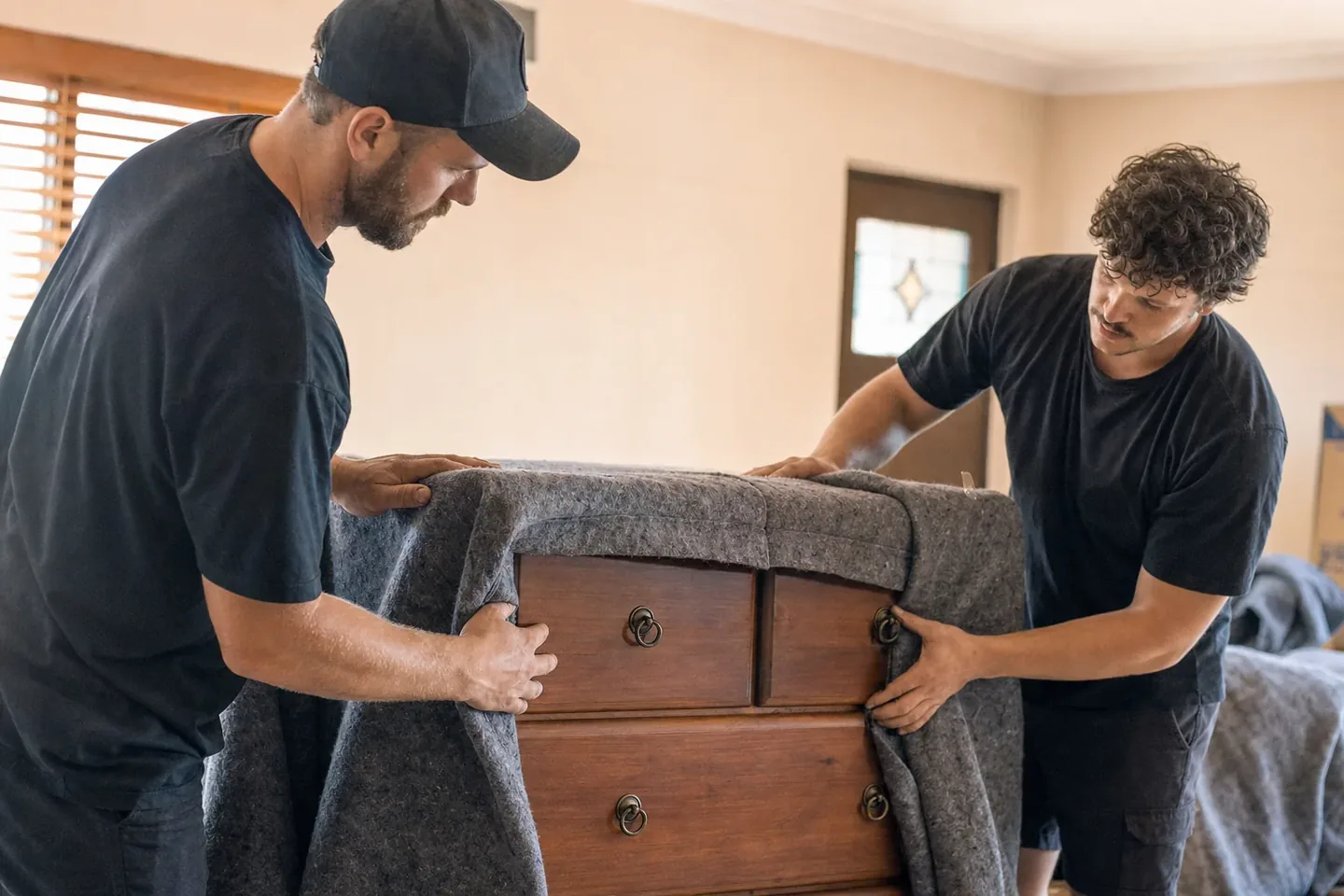 Two Perth removalists carefully wrapping a wooden chest of drawers in moving blankets